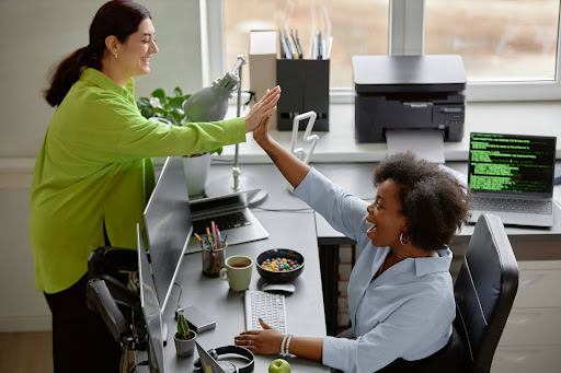 Two engaged employees celebrate a job well done in a collaborative workspace. A woman with light skin and dark hair, wearing a green shirt, leans over a desk with computer monitors to high-five her colleague, who has darker skin and curly hair, dressed in a blue dress shirt. In the background, natural light streams in through a window, illuminating a coding monitor, a printer, a filing cabinet, and a selection of healthy snacks. Their smiles reflect a positive workplace culture and a strong sense of employee appreciation and collaboration.