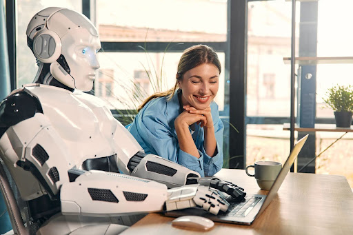 A robotic humanoid and a woman sit at a desk, seamlessly integrating AI in HR as they use a laptop together. The woman smiles, sipping from a coffee cup. The room is well-lit with large windows, fostering an atmosphere of innovation and employee satisfaction.