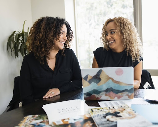 Two women smile at one another as though laughing. They are seated at a large table covered in magazines and reports, sharing a laptop with a case that looks like artistic mountains. Behind them is a window and a plant.