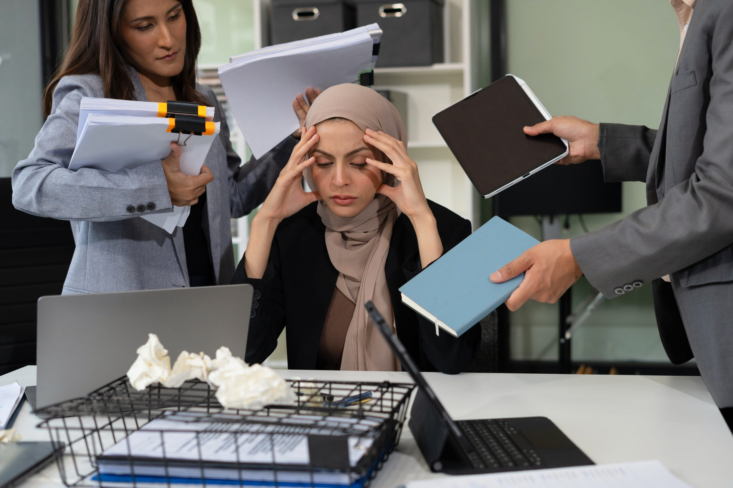 A woman in hijab at a desk, holding her head, while others engage in a conversation about HR outsourcing strategies.