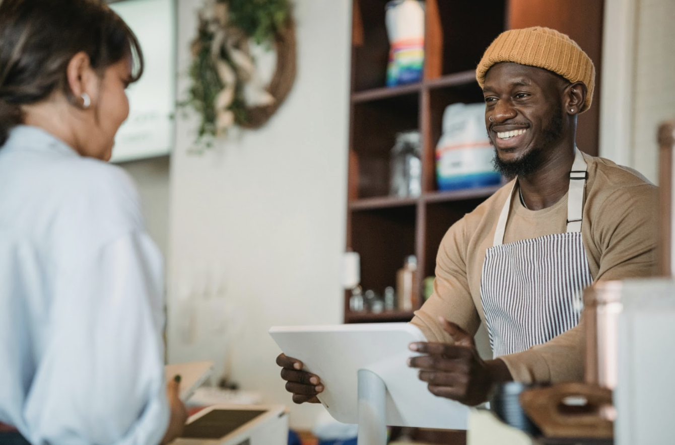 A man smiles while talking to a woman at a coffee shop, illustrating a warm human experience in a small business setting.