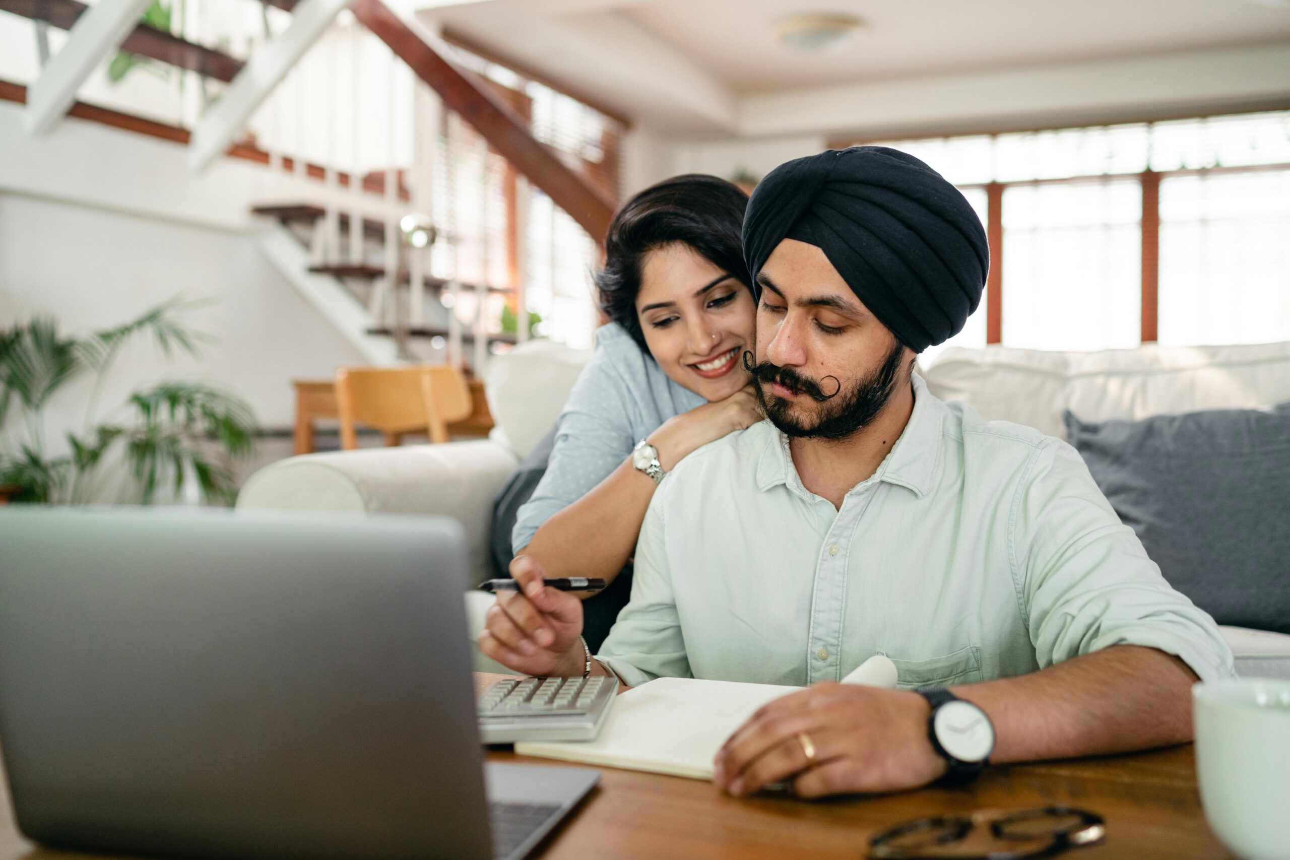 A man works remotely from a laptop and his wife stands behind him, smiling. They are in their home.